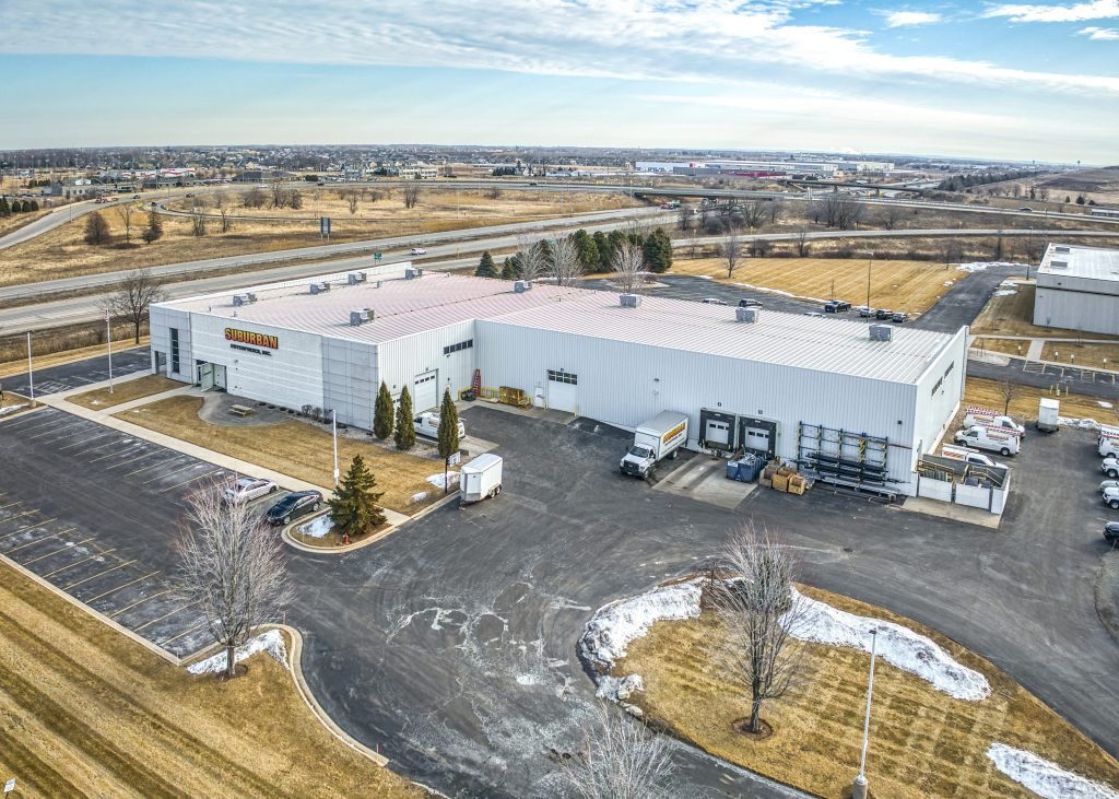Aerial view of a large industrial building with a parking lot, located near a highway, featuring the word "Suburban" prominently displayed on the facade. The image showcases the expansive layout, with delivery trucks and trees surrounding the facility.