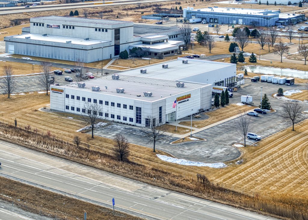 Aerial view of Suburban Manufacturing facility located near a highway, featuring a large gray building with a sign, surrounded by a paved parking lot and bare landscaping.