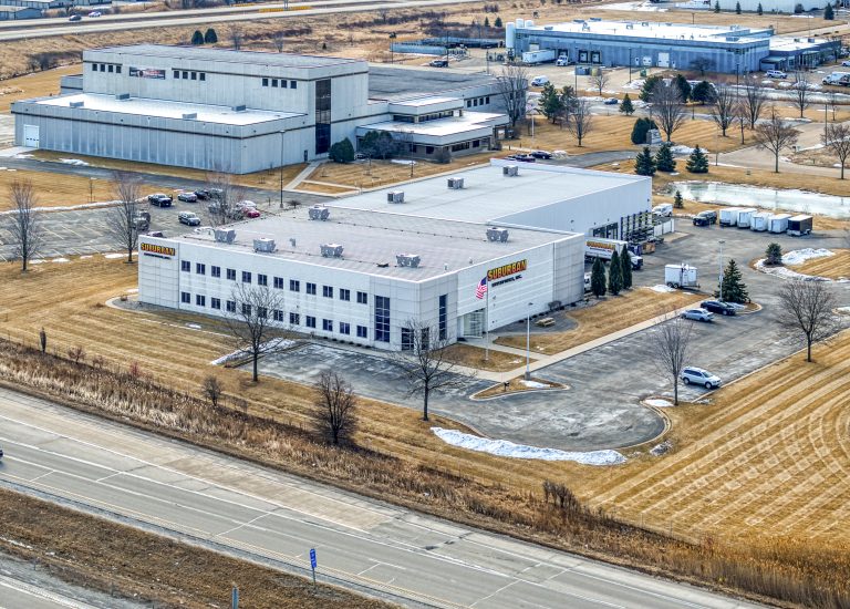 Aerial view of Suburban Manufacturing facility located near a highway, featuring a large gray building with a sign, surrounded by a paved parking lot and bare landscaping.
