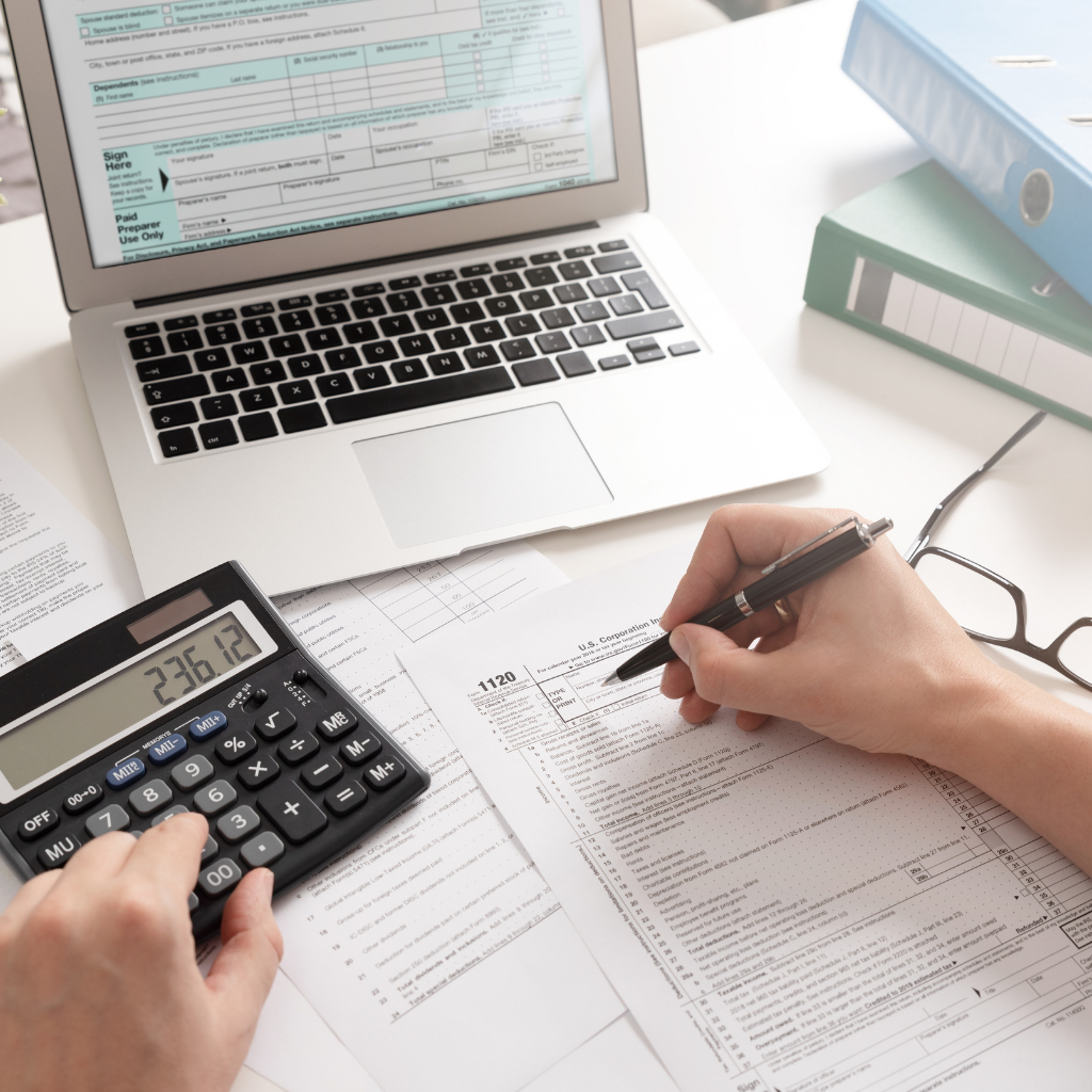 Person calculating taxes with a calculator while filling out tax forms on a desk with a laptop and organized paperwork in the background.