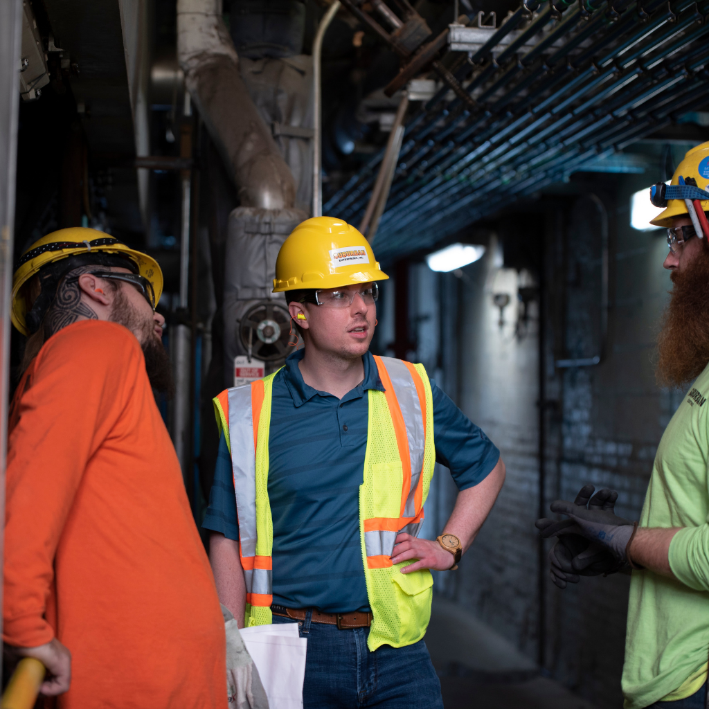 Construction workers in safety gear discussing project details in a dimly lit industrial environment. The image features three men: one in an orange shirt and a hard hat, another wearing a blue shirt with a reflective vest, and the third in a green shirt. They are surrounded by pipes and machinery, highlighting a collaborative work atmosphere.