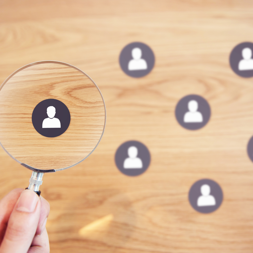 Hand holding a magnifying glass over a group of user icons on a wooden surface, symbolizing user research or talent identification.