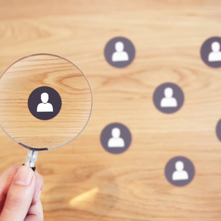 Hand holding a magnifying glass over a group of user icons on a wooden surface, symbolizing user research or talent identification.