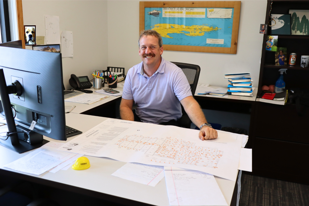A smiling man in a light purple polo shirt sits at a desk in an office, surrounded by documents, a computer monitor, and office supplies. He is pointing at a large printed plan on the desk that features various markings. In the background, there is a map on the wall and a stack of blue books.