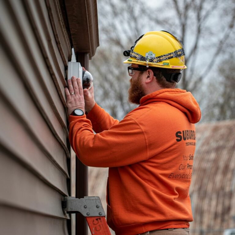Worker in a yellow hard hat and orange sweatshirt installing equipment on a wooden exterior wall, showcasing safety and precision in construction or maintenance tasks.
