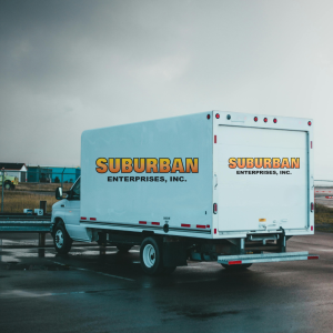 White delivery truck with "Suburban Enterprises, Inc." branding parked on a wet surface, showcasing the company's logo and a cloudy sky in the background.