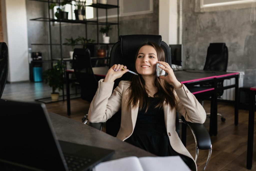 Young professional woman sitting in an office chair, smiling while talking on a smartphone and holding a pen, with a modern workspace in the background featuring desks and plants.